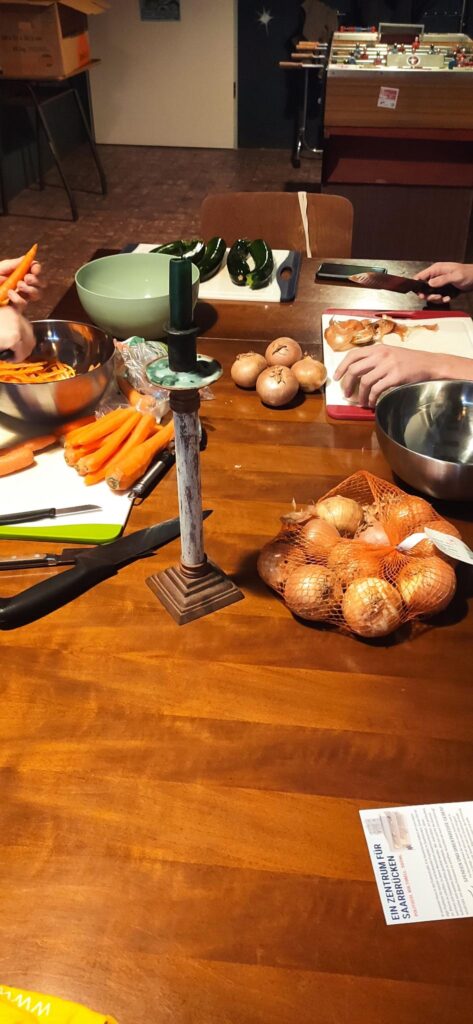 Zwiebeln und Karotten werden geschält und auf einem Schneidebrett geschnitten. Im Vordergrund steht ein Kerzenständer mit grüner Kerze, im Hintergrund ist ein Tischkicker zu erkennen.