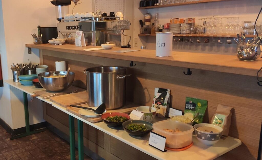 Eine Theke mit einer Spendendose darauf, davor eine Schüssel mit Spaghetti, ein großer Topf und verschiedene Toppings in Schälchen.